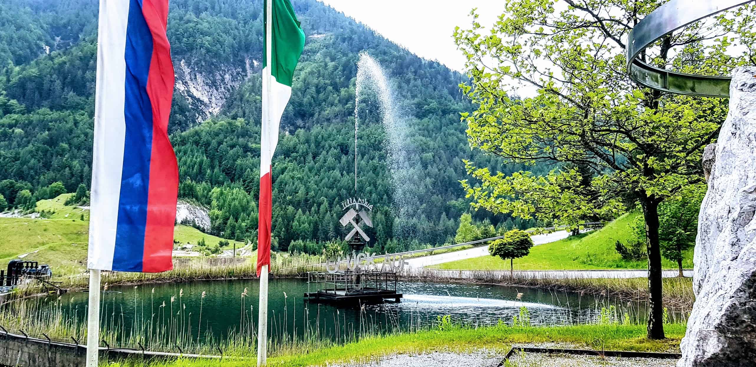 Teich mit Brunnen und Fahnen vor Schaubergwerk Terra Mystica im Naturpark Dobratsch